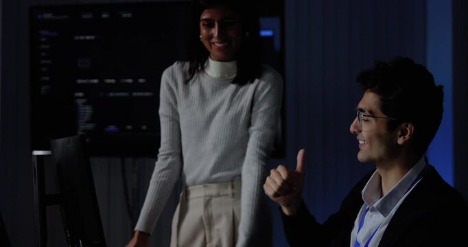 Adult female technology professional and seated male colleague smile, exchange high five, then separate after reviewing software dashboard, expressing successful teamwork during project workflow.
