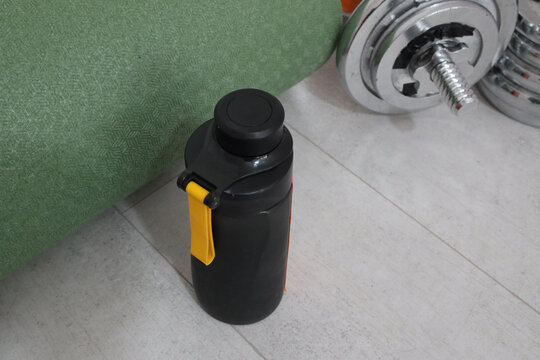 Black Insulated Water Bottle With Yellow Clip Stands Beside Chrome Dumbbell And Green Exercise Mat On Light Tiled Floor, Creating Simple Home Fitness Scene With Workout Gear, Training, And Hydration