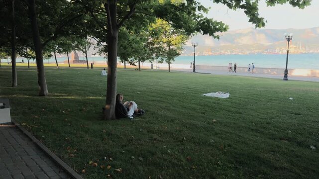 Unrecognizable young woman sitting on green grass under a tree reading a book in coastal city park with pedestrians walking on sea promenade and distant mountains during peaceful summer sunset.