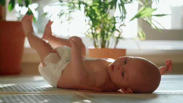Sweet newborn baby in a diaper resting on a play mat, bathed in gentle sunlight. The curious infant with blue eyes actively moves its little arms and legs, exploring the new world