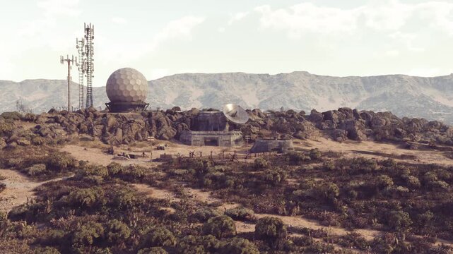 A communication facility with radar dishes and towers is situated on rocky terrain surrounded by sparse vegetation and mountains under a clear sky during the day.