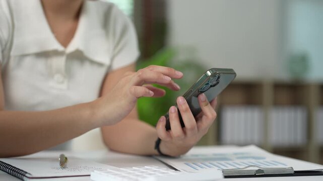 Businesswoman using a smartphone at her office desk with documents and a calculator, scrolling and typing on the screen to manage business and finances online