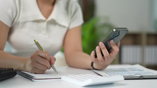 Businesswoman calculating financial data using a smartphone and taking notes while working at her desk in the office, managing company expenses and budget planning