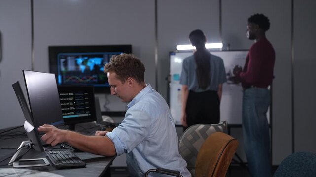 Caucasian male programmer coding on computer monitors in modern IT office, Software developer working on future technology business innovation while diverse colleagues brainstorming in background