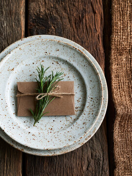 Rustic kraft paper gift wrapped with rosemary and twine on ceramic plate, creating warm natural still life with tactile textures and earthy styling