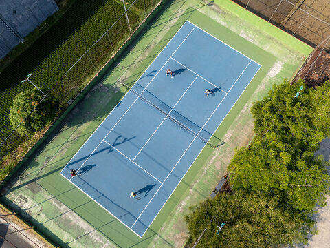 Aerial view of a blue tennis court with four people playing, surrounded by lush green trees and fields in Klaten, Central Java, Indonesia.