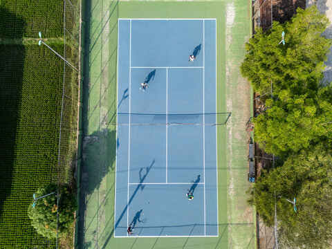 Aerial view of people playing tennis on a blue court surrounded by lush green fields and trees in Klaten, Central Java, Indonesia.