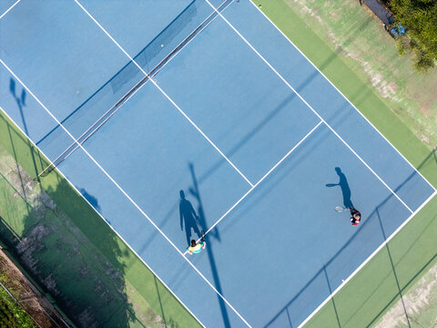 Aerial view of two people playing tennis on a blue court with long shadows cast by the surrounding floodlights in Klaten, Central Java, Indonesia.