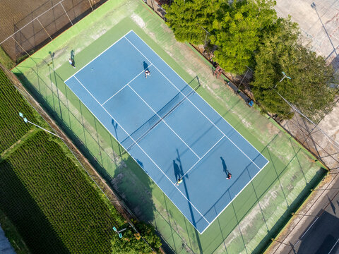 Aerial view of a blue tennis court with people playing, surrounded by green fields and trees in Klaten, Central Java, Indonesia.