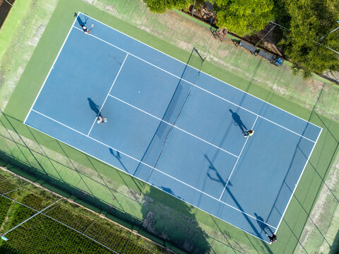 Aerial view of a blue tennis court with players in action, casting long shadows on the surface in Klaten, Central Java, Indonesia.