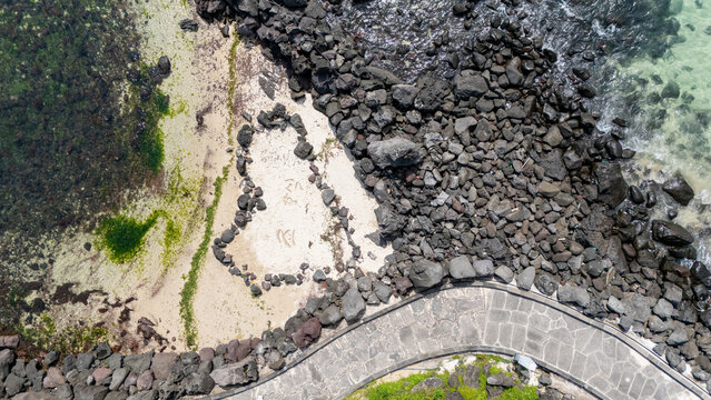 Aerial view of a heart-shaped arrangement of volcanic stones on a sandy beach next to a stone walkway and dark basalt rocks Jeju-si, Jeju-do, South Korea.