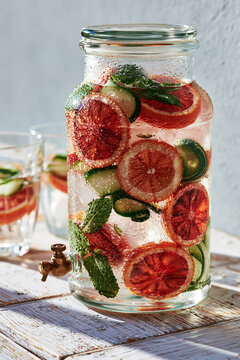 A large glass dispenser or pitcher filled with ice-cold water infused with slices of fresh blood orange, cucumber ribbons, and fresh mint leaves.