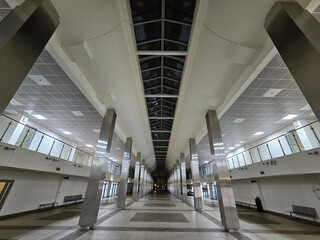 Fototapeta premium Empty station hall with tall columns and skylights. Ceiling panels and tiled floors reflect overhead lighting. Passenger benches line both sides of the corridor