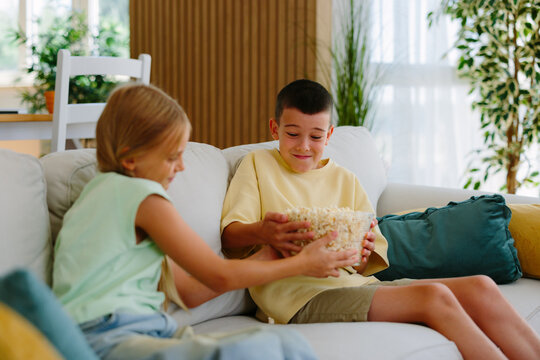 Two young siblings are sitting together on a white sofa in a bright living room, with a boy reluctantly sharing a bowl of popcorn with his sister, highlighting sibling rivalry over snacks
