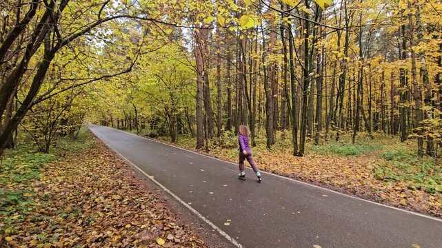 Fall foliage frames a paved path through a forest. Person skates down the path. They glide through trees with golden leaves. Leaves scatter along the ground. The scene is peaceful and active