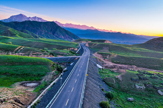 Scenic highway stretching through green rolling hills towards majestic mountain range under bright sunrise sky, ideal for travel and freedom concepts.