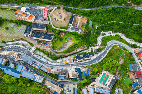 Top down view of a mountain settlement with winding asphalt roads, lush green vegetation, river, and residential buildings.
