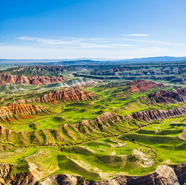 Panoramic high-angle view of colorful Danxia landforms and lush green pastures under a clear blue sky in Xinjiang, China.