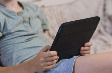 Young boy holding digital tablet sitting on couch at home. Child studying online, playing games or watching video content on modern portable device indoors. Technology and education concept. ( © Тетяна Хіміченко