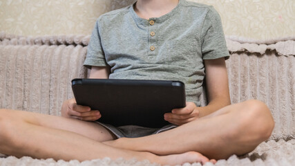 Young boy holding digital tablet sitting on couch at home. Child studying online, playing games or watching video content on modern portable device indoors. Technology and education concept. ( © Тетяна Хіміченко