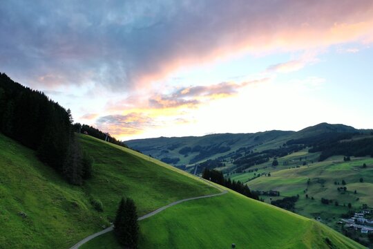 Aerial view of lush green mountain slopes at sunset, with a winding path through the grass and distant peaks under a colorful sky in Saalbach-Hinterglemm, Salzburg, Austria.