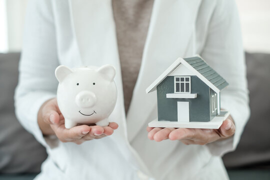 Woman holding white piggy bank and house model for home savings investment property purchase mortgage loan and financial planning concept