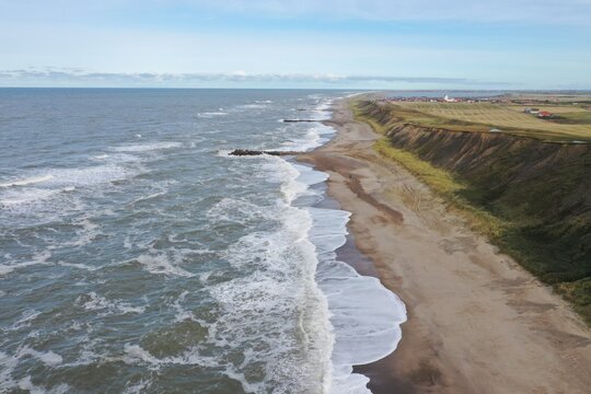 Aerial view of the coastline at Ferring, featuring dramatic cliffs, sandy beach, and waves crashing against stone groynes in Ferring, Skanderborg, Denmark.