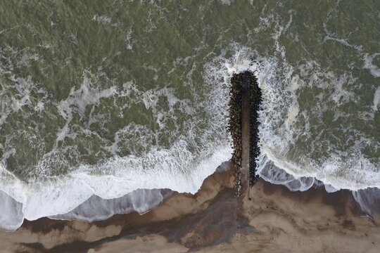 Aerial view of a stone groyne extending into the North Sea with waves crashing against the sandy beach in Ferring, Skanderborg, Denmark.