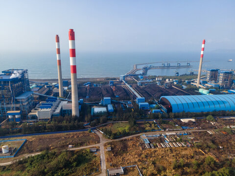 Aerial view of a large coal-fired power plant with tall red and white striped chimneys, industrial buildings, and coal storage facilities in Central Java, Indonesia.