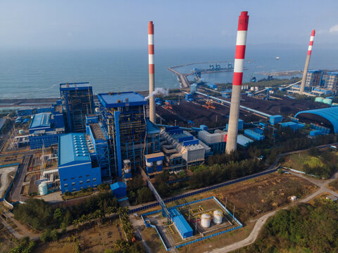 Aerial view of a coal-fired power plant with tall red-and-white striped chimneys, blue industrial buildings, and a coal storage yard along the coast, Central Java, Indonesia.