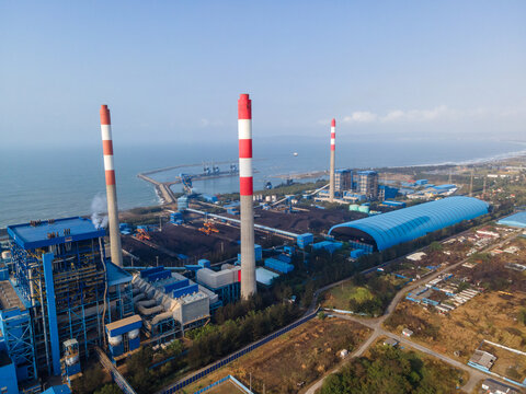 Aerial view of a large coal-fired power plant with tall red-and-white striped chimneys, industrial buildings, and coal storage facilities, Central Java, Indonesia.