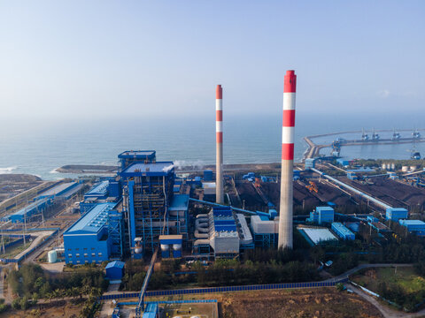Aerial view of a large coal-fired power plant with tall red-and-white striped chimneys and industrial buildings situated along the coast in Central Java, Indonesia.