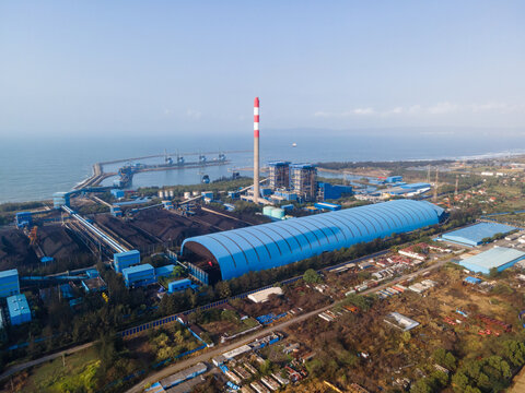 Aerial view of a large coal-fired power plant with a tall chimney and blue roof structures along the coast in Central Java, Indonesia.