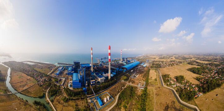 Aerial view of a large coal-fired power plant with tall chimneys and industrial facilities along the coastline, surrounded by rural fields and vegetation in Central Java, Indonesia.