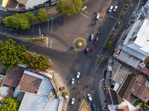 Aerial view of a busy roundabout intersection with cars and motorcycles navigating the roads surrounded by urban buildings and lush trees in Surakarta, Central Java, Indonesia.
