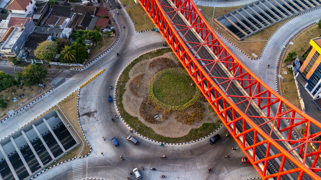 Aerial view of a prominent red steel railway bridge crossing over a busy urban roundabout with traffic and landscaped greenery in Banjarsari, Central Java, Indonesia.