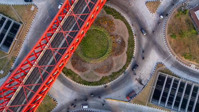 Aerial view of a striking red railway bridge crossing over a busy roundabout with traffic and a central landscaped garden in Banjarsari, Central Java, Indonesia.