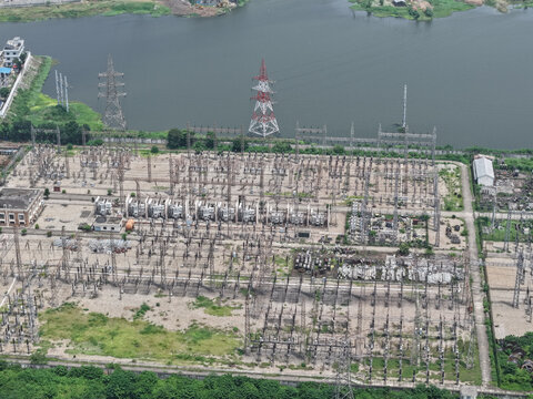 Aerial view of an electrical substation with power lines, transformers, and metal structures situated near a river in Dhaka, Dhaka Division, Bangladesh.