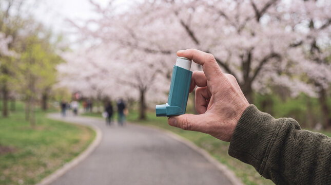 Hand holding a teal asthma inhaler against a blurred cherry blossom background