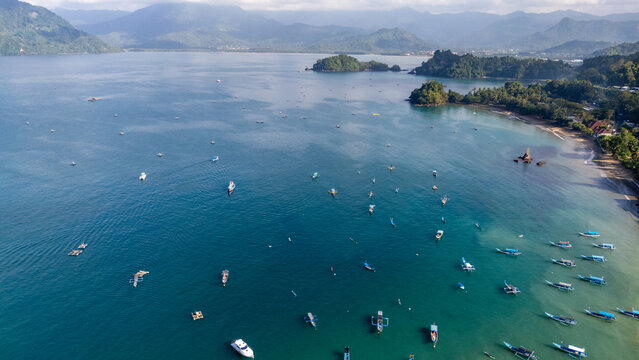 Aerial view of Pantai Karanggongso with numerous small fishing boats anchored in the clear blue bay, surrounded by lush coastal hills in Watulimo, East Java, Indonesia.