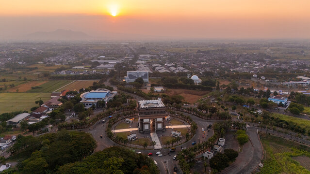 Aerial view of the Simpang Lima Gumul monument at sunset, surrounded by lush green fields and urban development in Kediri, East Java, Indonesia.