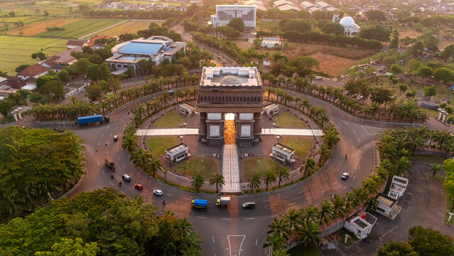 Aerial view of Simpang Lima Gumul Monument surrounded by a circular road, lush palm trees, and agricultural fields in the golden hour light in Kediri, East Java, Indonesia.
