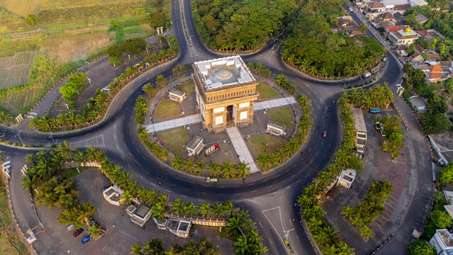 Aerial view of the Simpang Lima Gumul monument situated in the center of a large roundabout, surrounded by palm trees and urban infrastructure in Kediri, East Java, Indonesia.