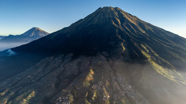 Aerial view of Mount Sumbing with terraced agricultural fields on its slopes and a small village at the base under a clear blue sky in Kaliangkrik, Central Java, Indonesia.