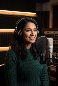 Smiling young woman wearing black headphones while recording professional audio in a dark cozy music studio