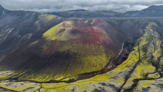 Aerial view of Raudaskal crater featuring vibrant red volcanic soil and bright green moss-covered slopes in the rugged landscape of Hella, Rangarthing ytra, Iceland.