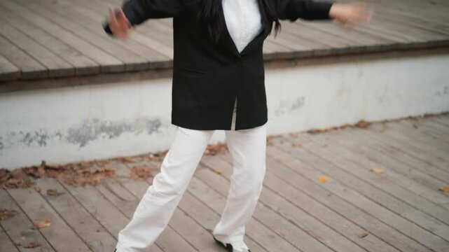 Close up of woman performing slow dance movements on outdoor wooden deck with scattered leaves. Woman wears white pants and black top moving gracefully through various poses