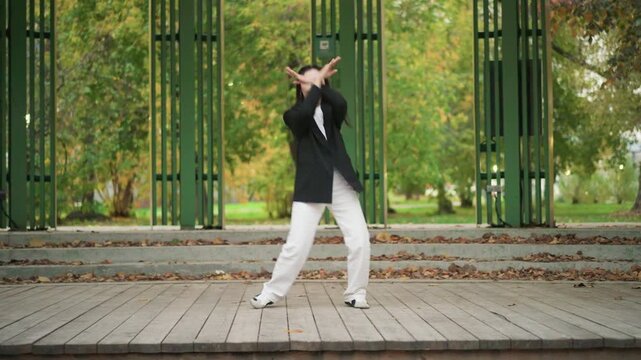Young woman performs dance routine on outdoor wooden stage with green metal railings surrounded by autumn foliage. Woman moves gracefully through various poses and gestures in park setting