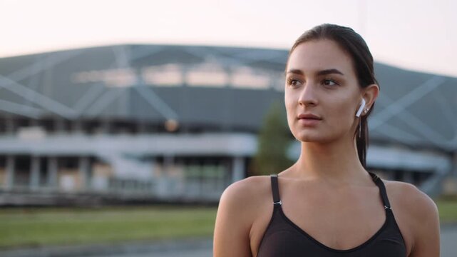 Close three-quarter portrait of young woman in black athletic top wearing white wireless earbud, standing outdoors in front of large modern arena with geometric facade and horizontal openings