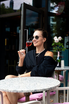 Stylish woman in sunglasses enjoys rose champagne at outdoor cafe, smiling while holding elegant glass near flowers. Relaxed summer autumn portrait leisure, luxury, fashion, travel, urban lifestyle